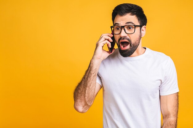 Jonge blanke man boos, gefrustreerd en woedend met zijn mobiele telefoon, boos op klantenservice. Geïsoleerd op gele achtergrond.