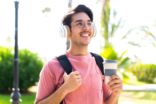 Jonge blanke man buiten in een park die muziek luistert en een afhaalkoffie houdt