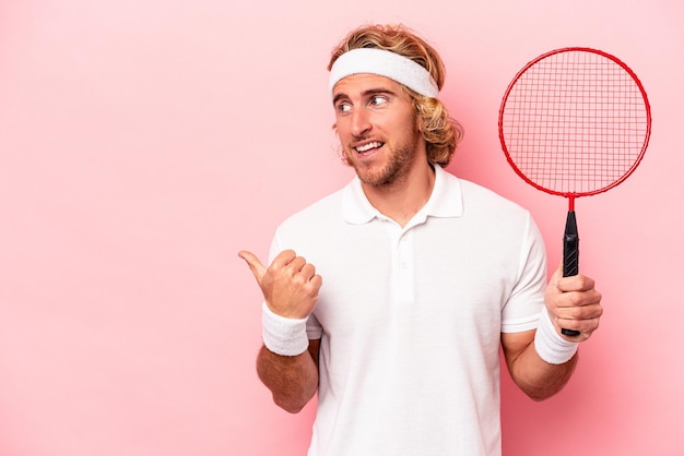 Jonge blanke man die badminton speelt geïsoleerd op roze achtergrondpunten met duimvinger weg, lachend en zorgeloos.