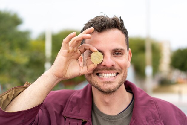 Jonge blanke man die buiten een Bitcoin vasthoudt met een gelukkige uitdrukking