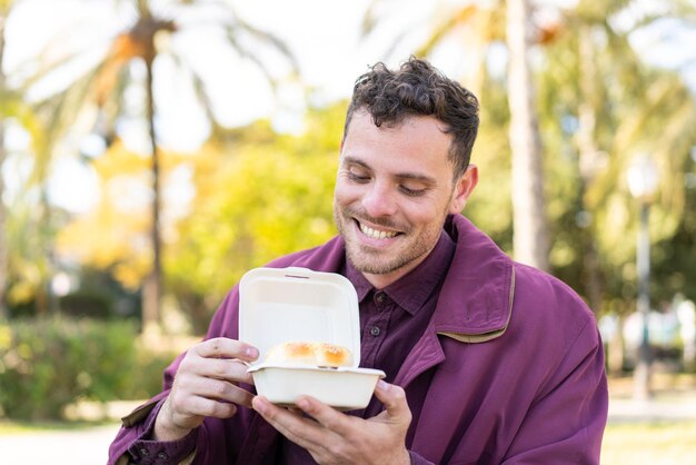 Jonge blanke man die buiten een hamburger vasthoudt met een gelukkige uitdrukking