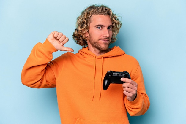 Jonge blanke man die speelt met een videogamecontroller geïsoleerd op een blauwe achtergrond voelt zich trots en zelfverzekerd, een voorbeeld om te volgen.