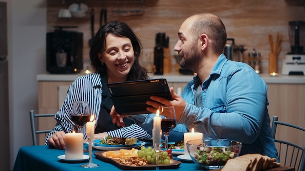 Jonge blanke man die tablet gebruikt tijdens een feestelijke maaltijd die aan zijn vrouw vakantiebestemmingen laat zien. Volwassenen die aan tafel zitten, online browsen, surfen, smartphones gebruiken, genieten van de maaltijd