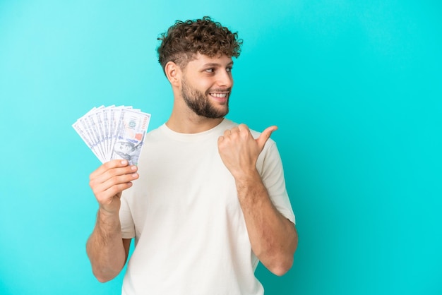 Jonge blanke man die veel geld neemt geïsoleerd op een blauwe achtergrond en naar de zijkant wijst om een product te presenteren