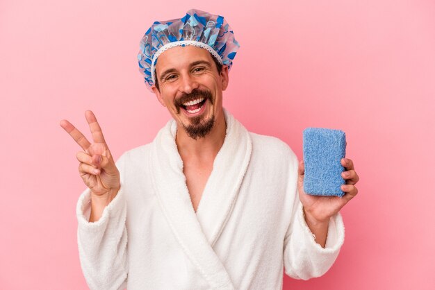 Jonge blanke man gaat naar de douche met spons geïsoleerd op roze achtergrond vrolijk en zorgeloos met een vredessymbool met vingers.