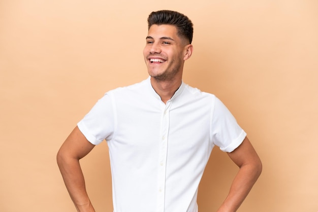 Jonge blanke man geïsoleerd op beige achtergrond poseren met armen op heup en lachend