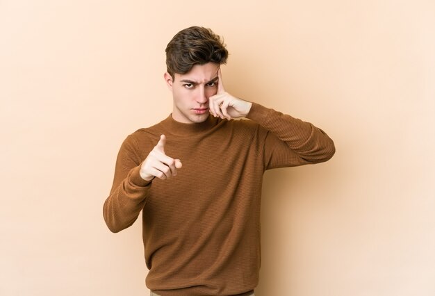 Jonge blanke man geïsoleerd op beige muur wijzende tempel met vinger, denken, gericht op een taak.