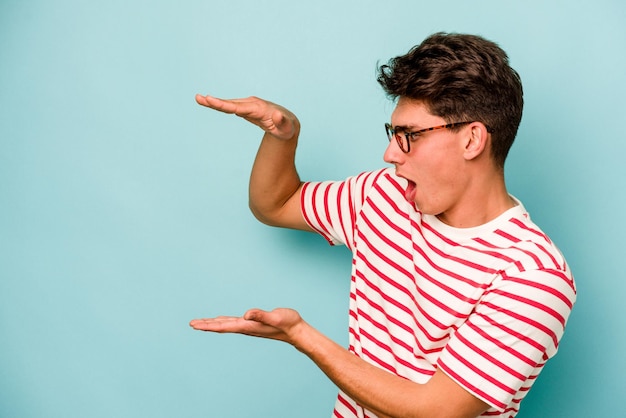 Jonge blanke man geïsoleerd op blauwe achtergrond geschokt en verbaasd met een kopie ruimte tussen handen