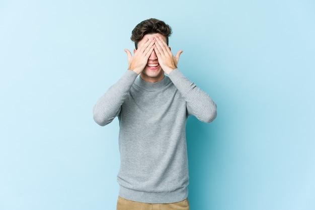 Jonge blanke man geïsoleerd op blauwe achtergrond heeft betrekking op ogen met handen, glimlacht in het algemeen wachtend op een verrassing.