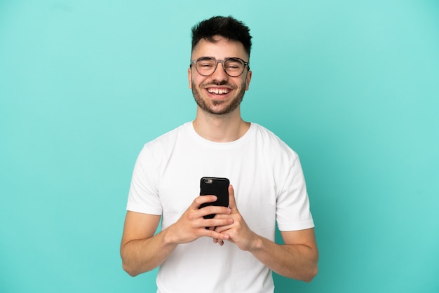 Jonge blanke man geïsoleerd op blauwe achtergrond kijken naar de camera en glimlachen tijdens het gebruik van de mobiel