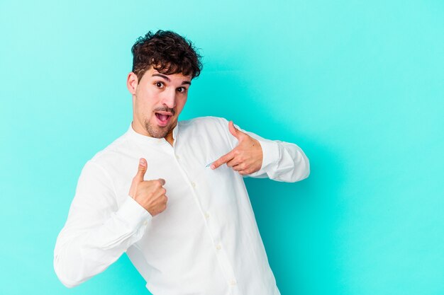 Jonge blanke man geïsoleerd op blauwe achtergrond verrast wijzend met de vinger, breed glimlachend.