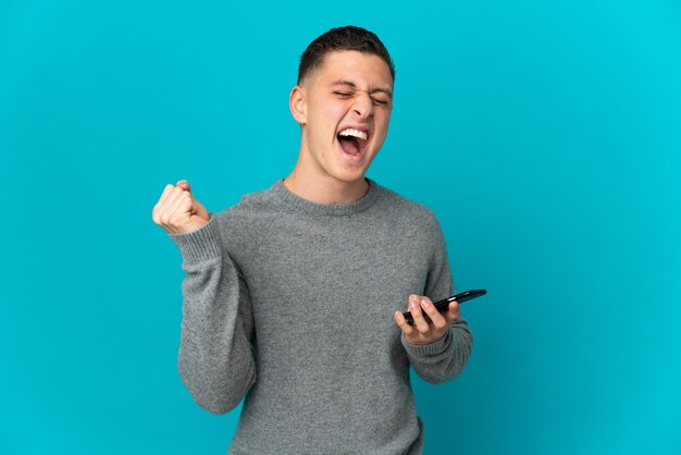 Jonge blanke man geïsoleerd op blauwe muur met behulp van mobiele telefoon en doet overwinningsgebaar