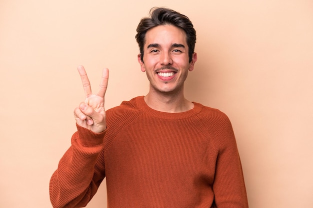 Jonge blanke man geïsoleerd op een beige achtergrond die overwinningsteken toont en breed glimlacht.