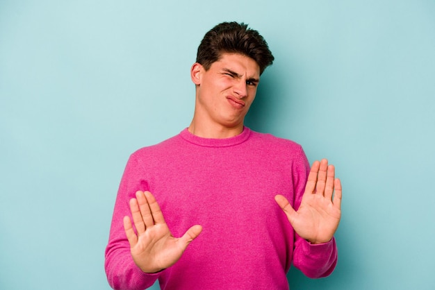Jonge blanke man geïsoleerd op een blauwe achtergrond die iemand afwijst die een gebaar van walging toont