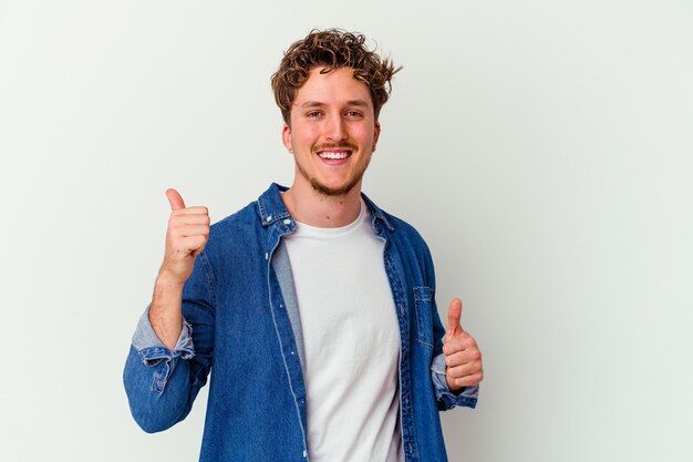 Jonge blanke man geïsoleerd op een witte muur verhogen beide duimen omhoog, glimlachend en zelfverzekerd.