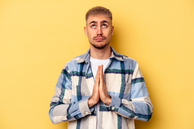 Jonge blanke man geïsoleerd op gele achtergrond hand in hand in bidden in de buurt van mond, voelt zich zelfverzekerd.