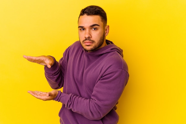 Jonge blanke man geïsoleerd op gele achtergrond met iets met beide handen, productpresentatie.