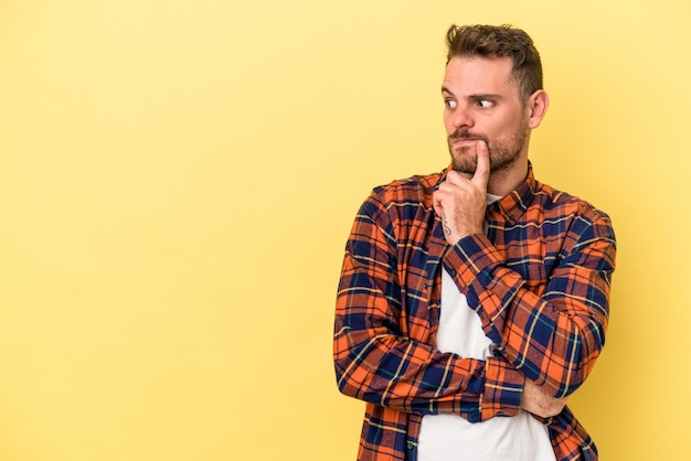 Jonge blanke man geïsoleerd op gele achtergrond zijwaarts kijkend met twijfelachtige en sceptische uitdrukking.