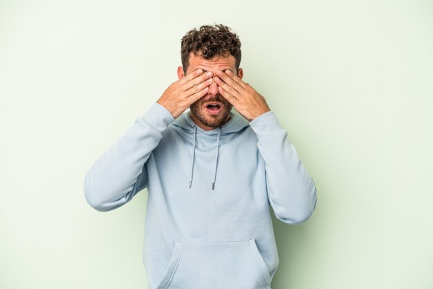 Jonge blanke man geïsoleerd op groene achtergrond bang voor ogen met handen.