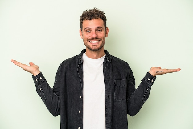 Jonge blanke man geïsoleerd op groene achtergrond maakt schaal met armen, voelt zich gelukkig en zelfverzekerd.