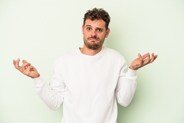 Jonge blanke man geïsoleerd op groene achtergrond twijfelen en schouders ophalen in ondervraging gebaar.