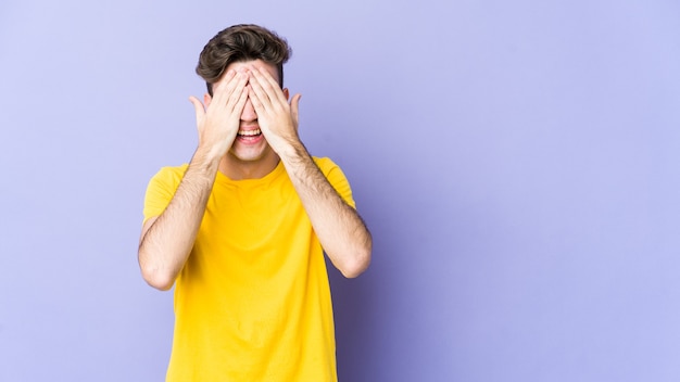 Jonge blanke man geïsoleerd op paarse ruimte bedekt ogen met handen, glimlacht in het algemeen wachtend op een verrassing.