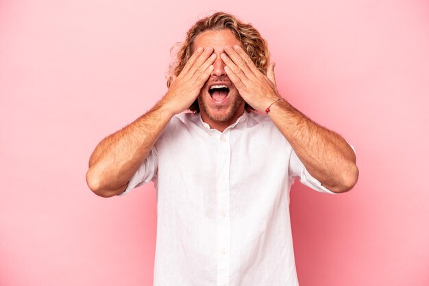 Jonge blanke man geïsoleerd op roze achtergrond bedekt ogen met handen, glimlacht in grote lijnen wachtend op een verrassing.