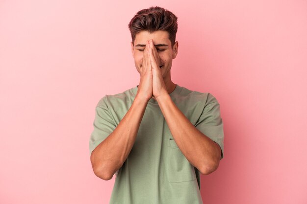 Jonge blanke man geïsoleerd op roze achtergrond hand in hand in bidden in de buurt van mond, voelt zich zelfverzekerd.