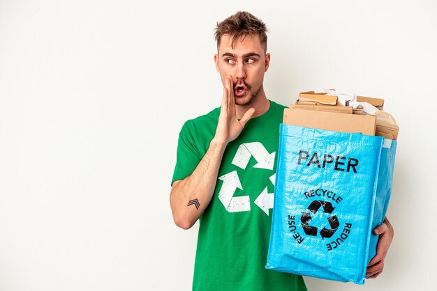 Jonge blanke man gerecycled papier geïsoleerd op een witte achtergrond zegt een geheim heet remnieuws en kijkt opzij