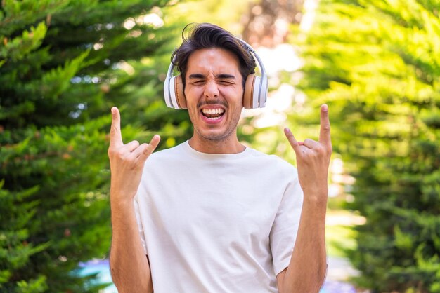 Jonge blanke man in een park die muziek luistert en rockgebaar maakt