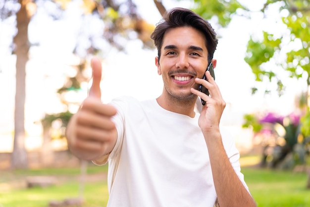 Jonge blanke man in een park met behulp van mobiele telefoon terwijl hij duimen omhoog doet