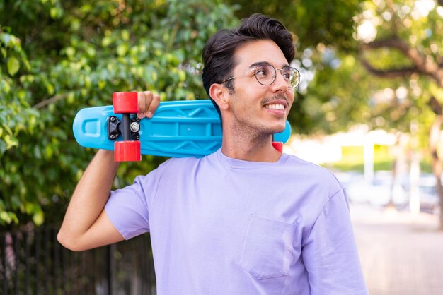 Jonge blanke man in een park met een skate met een gelukkige uitdrukking