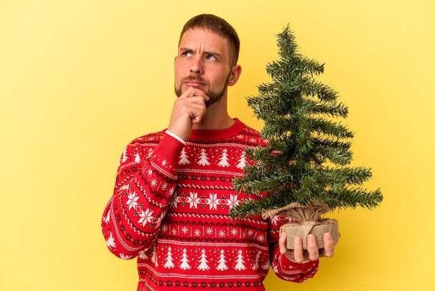 Jonge blanke man koopt kleine boom voor Kerstmis geïsoleerd op gele achtergrond en kijkt zijwaarts met twijfelachtige en sceptische uitdrukking.