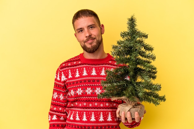 Jonge blanke man koopt kleine boom voor Kerstmis geïsoleerd op gele achtergrond gelukkig, glimlachend en vrolijk.