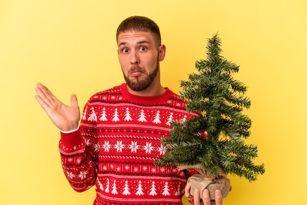 Jonge blanke man koopt kleine boom voor Kerstmis geïsoleerd op gele achtergrond haalt schouders op en open ogen verward.