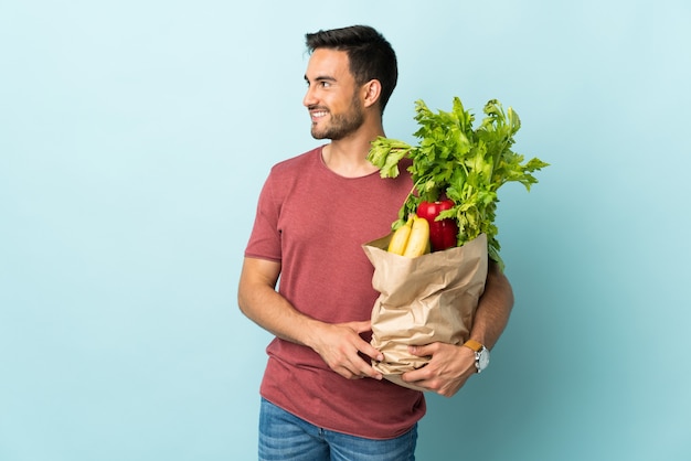 Jonge blanke man kopen wat groenten geïsoleerd op blauwe achtergrond op zoek naar de zijkant