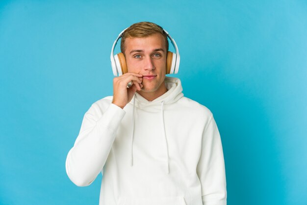 Jonge blanke man luisteren muziek geïsoleerd op blauwe muur met vingers op lippen een geheim houden.