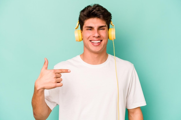 Jonge blanke man luisteren naar muziek geïsoleerd op blauwe achtergrond persoon die met de hand wijst naar een shirt kopieerruimte trots en zelfverzekerd?