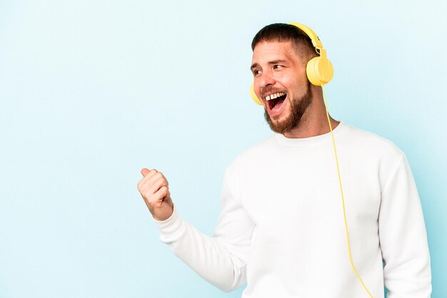 Jonge blanke man luisteren naar muziek geïsoleerd op blauwe achtergrond wijst met duimvinger weg, lachend en zorgeloos.