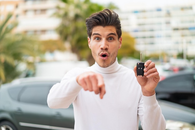 Jonge blanke man met autosleutels buitenshuis verrast en wijzend naar voren