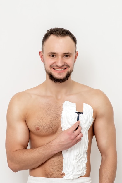 Jonge blanke man met baard houdt scheermes zijn borst met wit scheerschuim op witte muur. Man zijn torso scheren