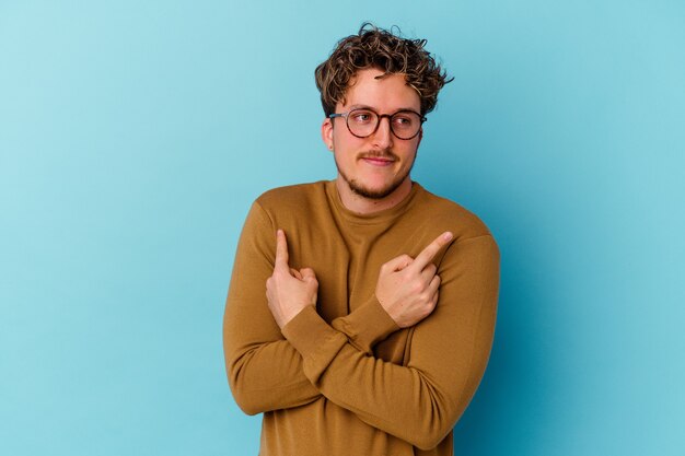 Foto jonge blanke man met bril geïsoleerd op blauwe achtergrond punten zijwaarts, probeert te kiezen tussen twee opties.