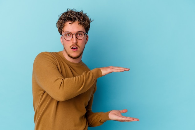 Jonge blanke man met bril geïsoleerd op blauwe muur geschokt en verbaasd met een kopie ruimte tussen handen.