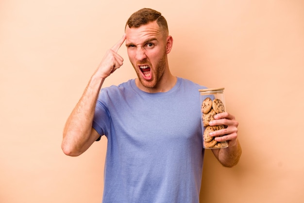 Jonge blanke man met cookies pot geïsoleerd op beige achtergrond met een teleurstelling gebaar met wijsvinger