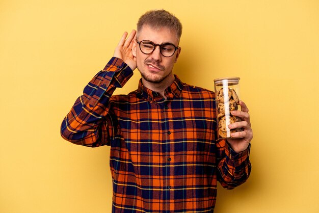 Jonge blanke man met cookies pot geïsoleerd op gele achtergrond probeert te luisteren naar een roddel.