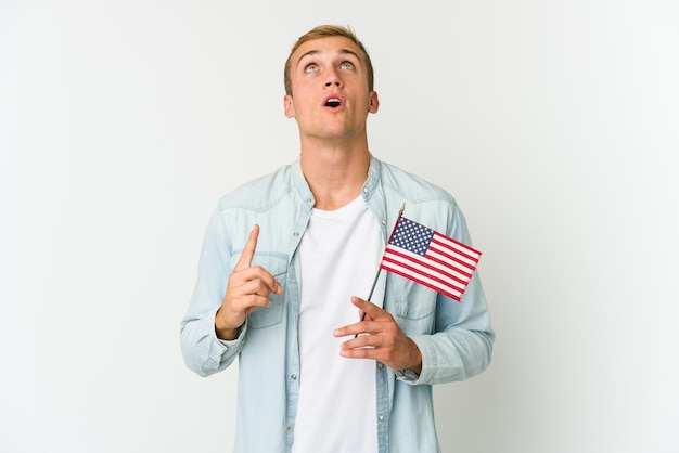 Jonge blanke man met een Amerikaanse vlag geïsoleerd op een witte muur naar boven gericht met geopende mond.