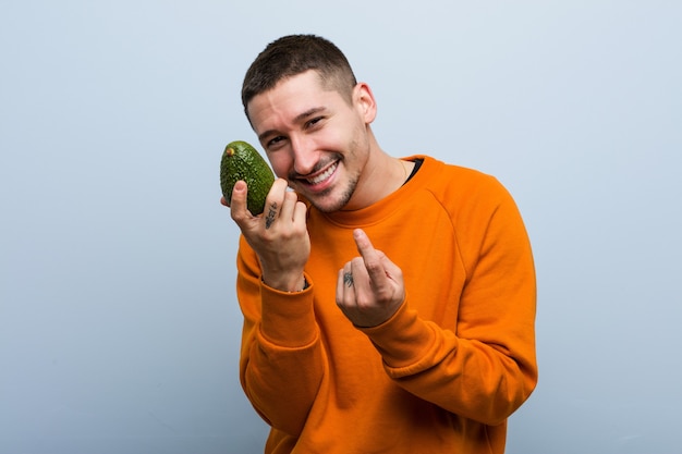 Jonge blanke man met een avocado die met de vinger naar je wijst alsof ze dichterbij komen.