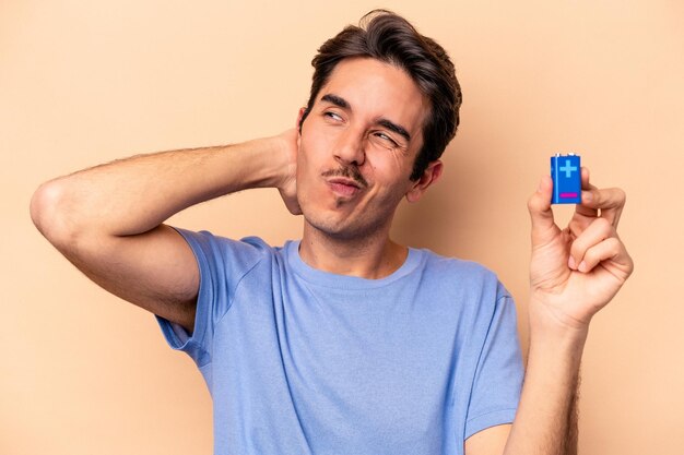 Jonge blanke man met een batterij geïsoleerd op een beige achtergrond die de achterkant van het hoofd aanraakt, denkt en een keuze maakt.