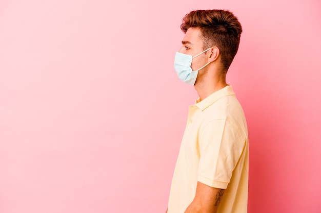 Jonge blanke man met een bescherming tegen coronavirus geïsoleerd op roze muur starend naar links, zijwaarts pose.