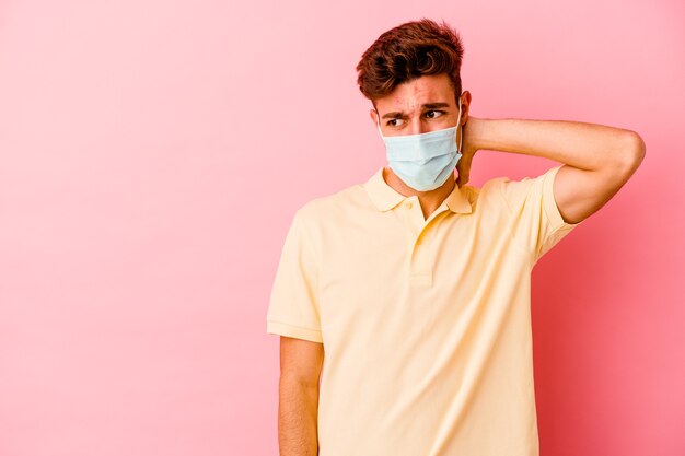 Jonge blanke man met een bescherming voor coronavirus geïsoleerd op een roze achtergrond die de achterkant van het hoofd aanraakt, denkt en een keuze maakt.